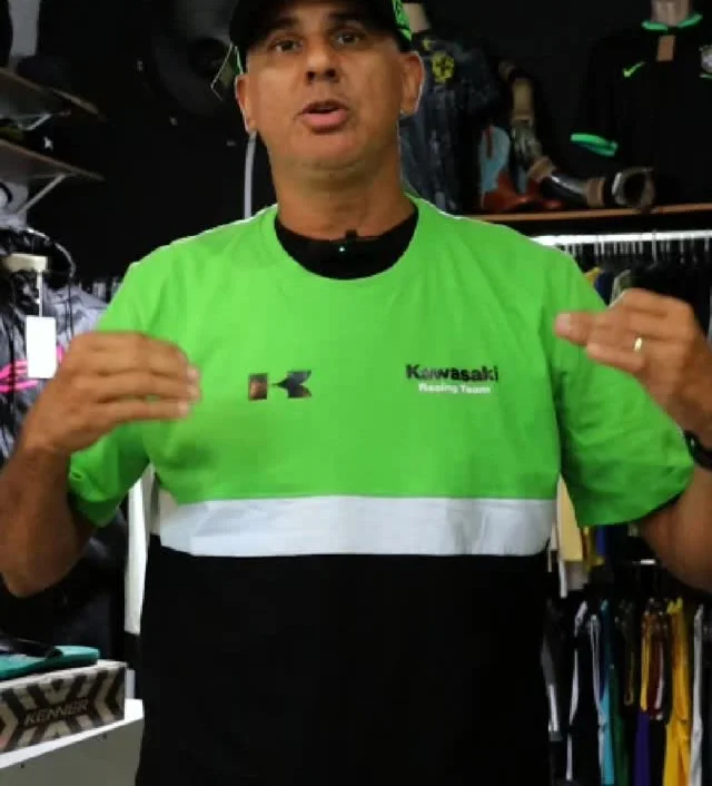 Angelo, dono da AGS Outlet, de boné e camisa verde, falando na frente da arara de camisas da loja